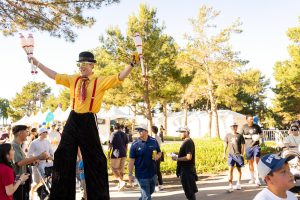 A stilt walker juggler waves hands up as attendees walk by
